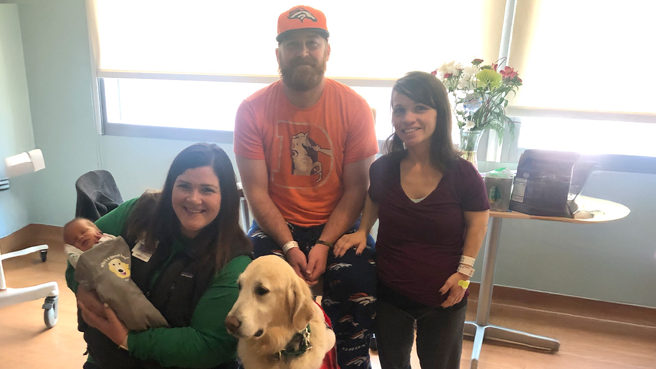 Three adults and a golden retriever in a hospital room, one adult kneeling while holding a baby and the dog, with the other two adults standing beside them.