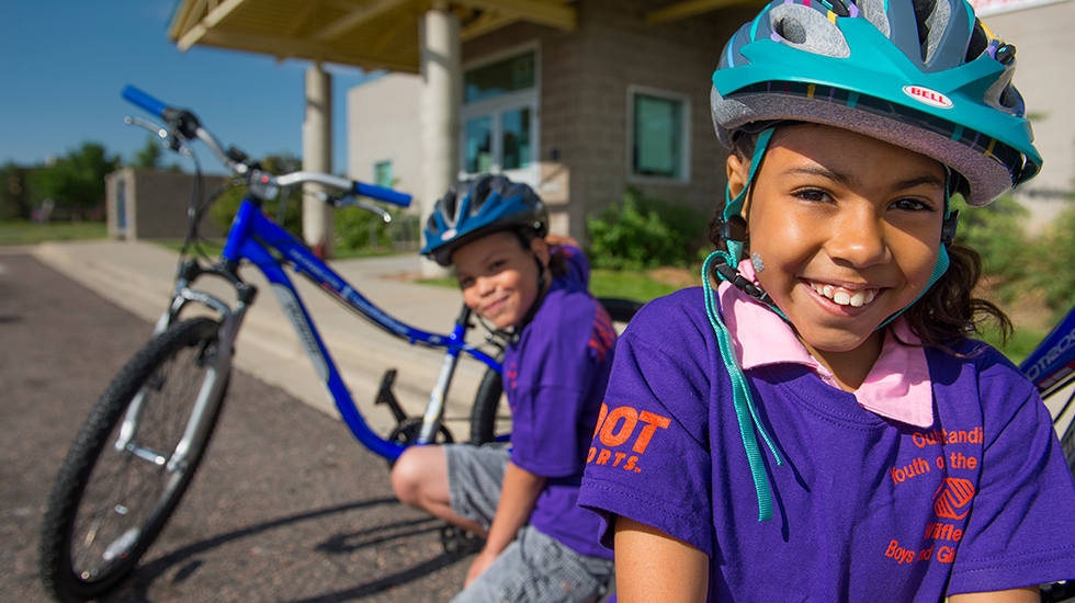 Two kids in purple shirts and blue bike helmets squat down next to their blue bikes outside of a brick building.