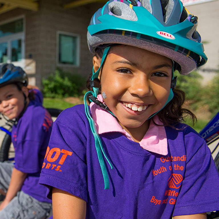 Two kids in purple shirts and blue bike helmets squat down next to their blue bikes outside of a brick building.