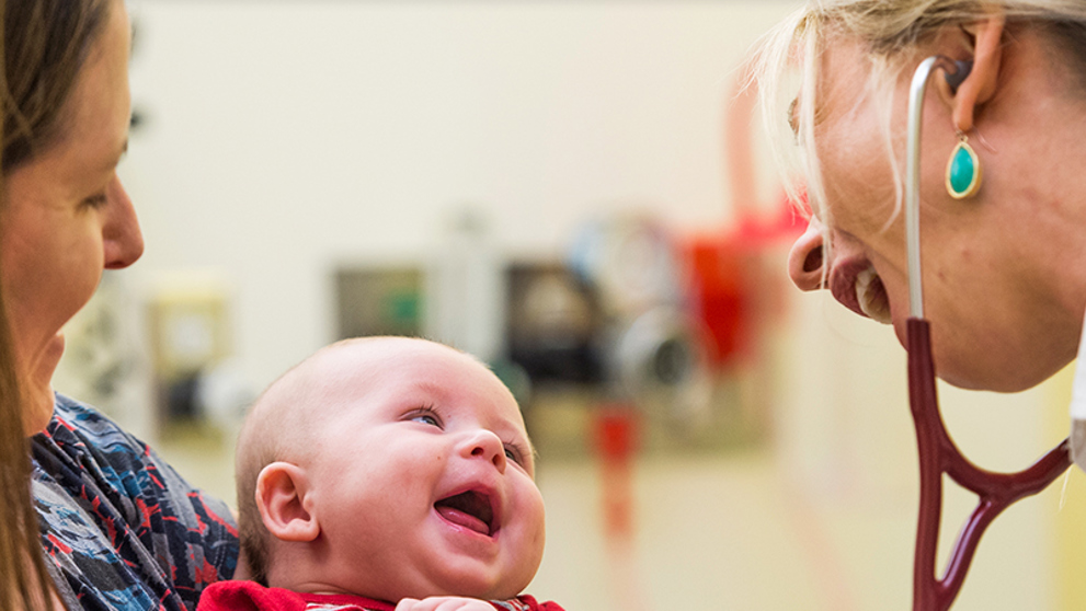 A woman with blonde hair in a ponytail is wearing a white lab coat and stethoscope while smiling at a baby being held by a woman with long brown hair.