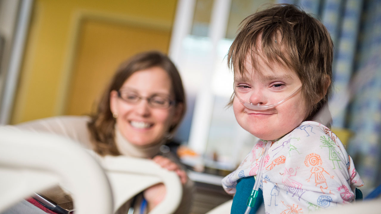 A patient with brown hair and nasal tubes sits in a hospital bed while a woman with long brown hair and glasses looks on from the background.