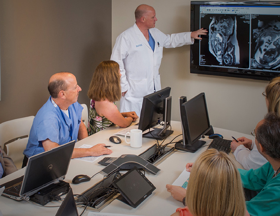 A group of doctors sit around a table with laptops while another doctor in a white coat stands next to a screen pointing to medical images.