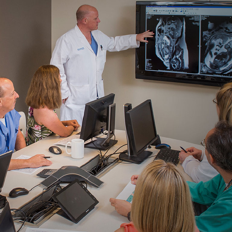 A group of doctors sit around a table with laptops while another doctor in a white coat stands next to a screen pointing to medical images.