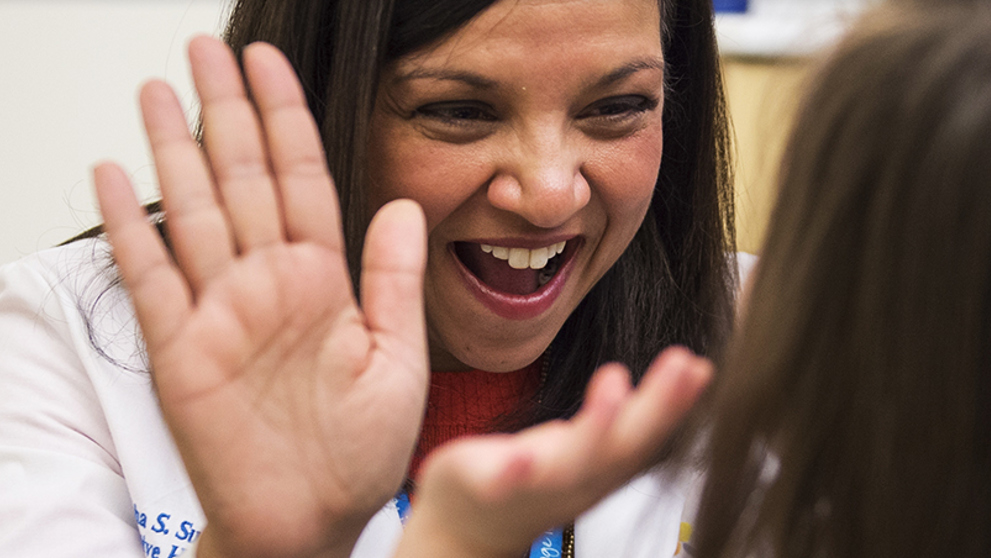 A close-up of Dr. Shika Sundaram, with long brown hair and wearing a white lab coat, high-fiving a patient with long brown hair.
