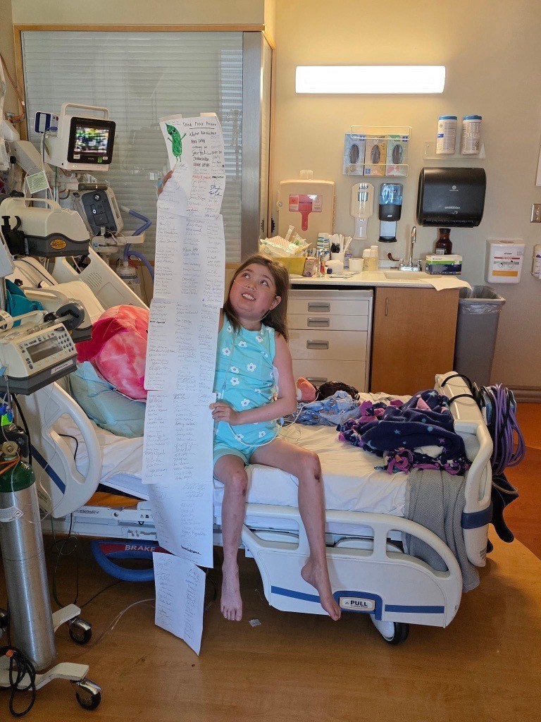 A young girl in a hospital bed holds up a long sheet of paper with signatures