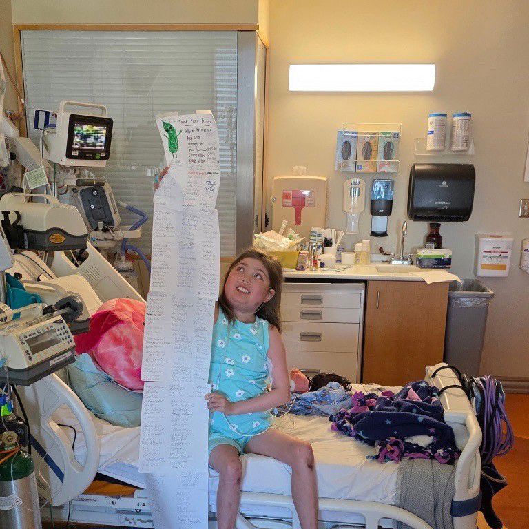 A young girl in a hospital bed holds up a long sheet of paper with signatures