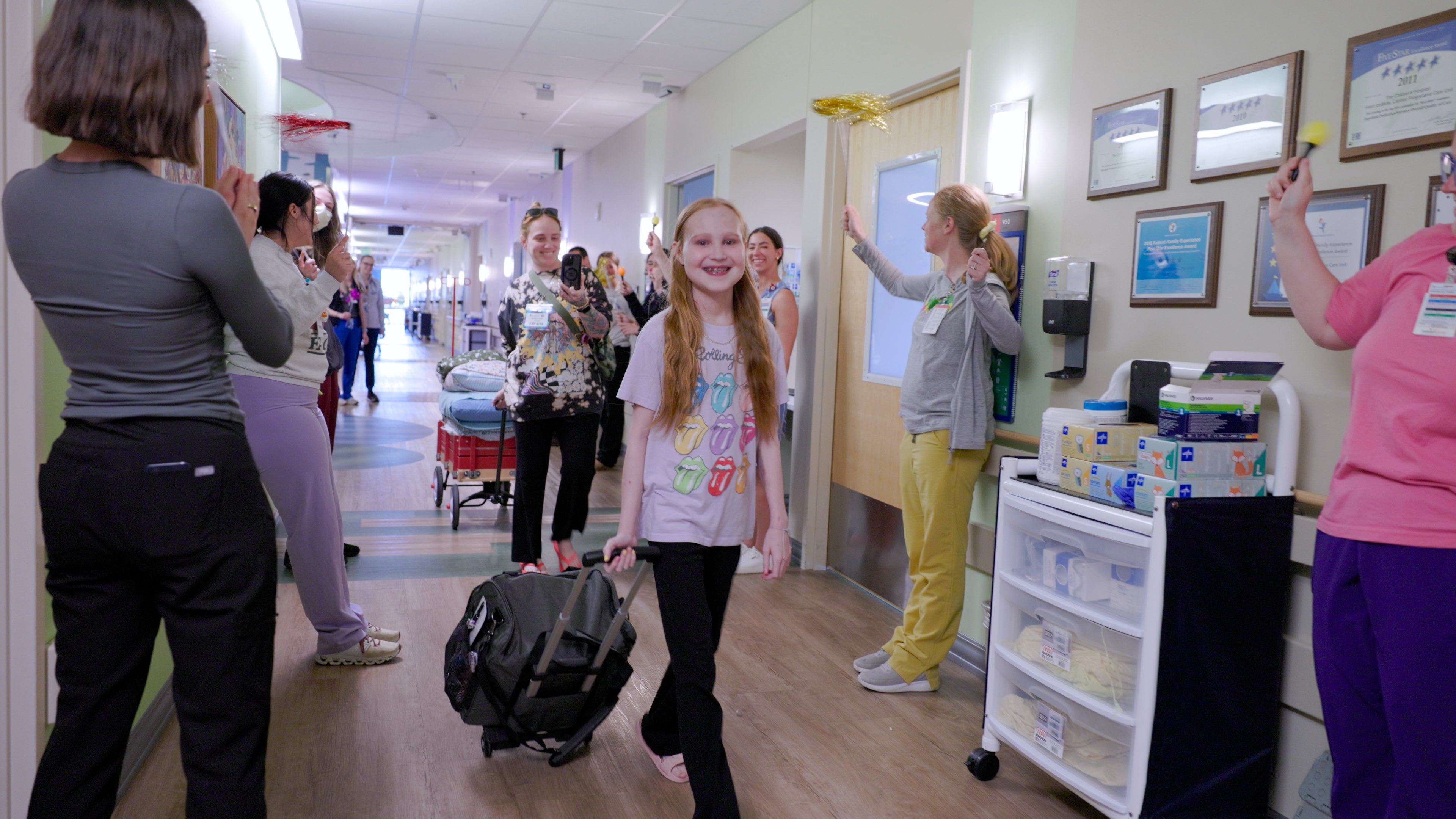 A young girl walks down a hospital hallway as staff members cheer. 