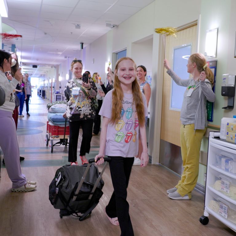A young girl walks down a hospital hallway as staff members cheer.