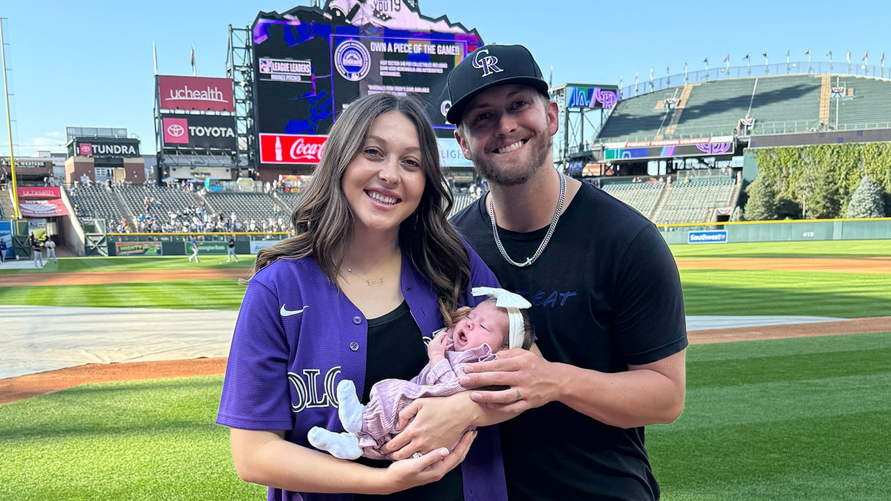 A couple smiles holding their young infant at a Rockies game