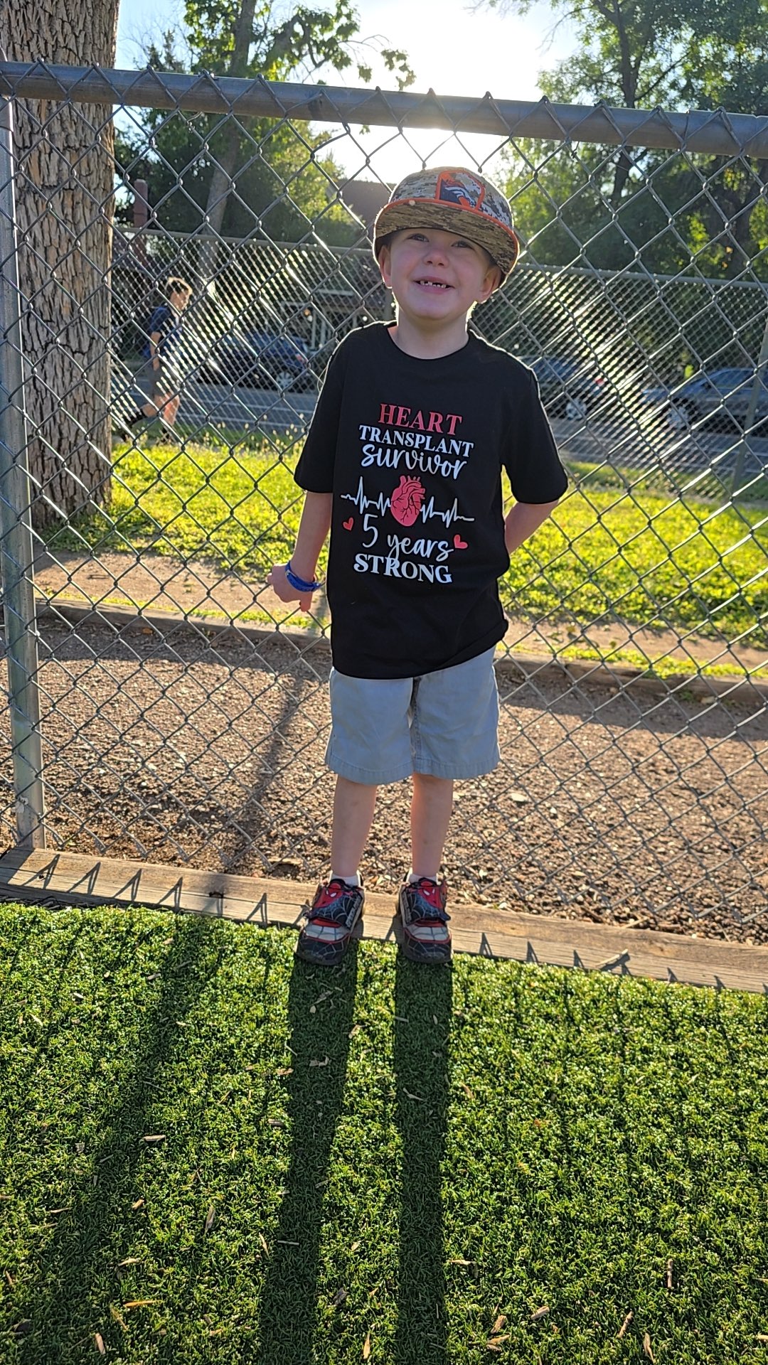 A young boy stands against a fence smiling. His shirt says heart transplant survivor 5 years strong
