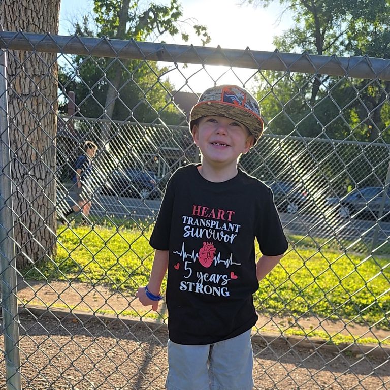 A young boy stands against a fence smiling. His shirt says heart transplant survivor 5 years strong