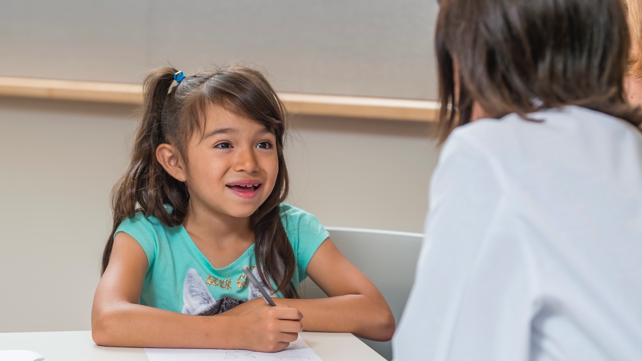 Girl with brown hair wearing a teal shirt is writing on a notepad and smiling at a doctor.
