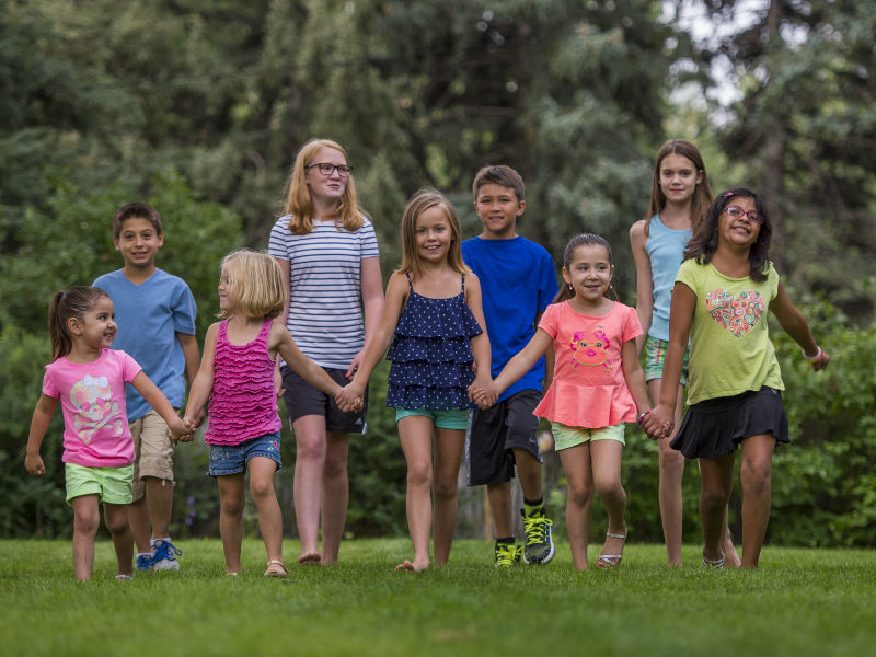 Pediatric Mental Health Impacting The Community A group of 9 kids - five in front and four in back - hold hands and walk in a grassy field with trees behind them. The kids are ages 4-15 and wearing brightly colored clothes in a variety of colors.