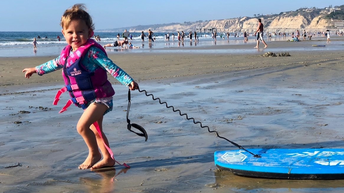 Emma smiles and pulls a boogie board behind her on the beach.