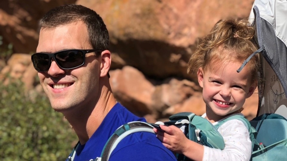 Emma and her dad smile while she rides in a hiking backpack that her dad carries.