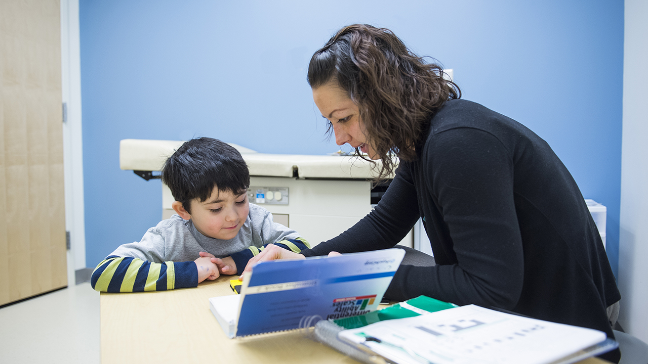 Intellectual Disability A boy wearing a blue and yellow striped shirt with a gray short sleeve shirt over it listens to a woman with short brown hair and wearing a black cardigan.