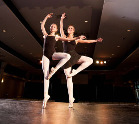 Two ballerinas in black leotards and white tights stand on one toe with the other foot held up by the knee, one arm up and one arm to the side.