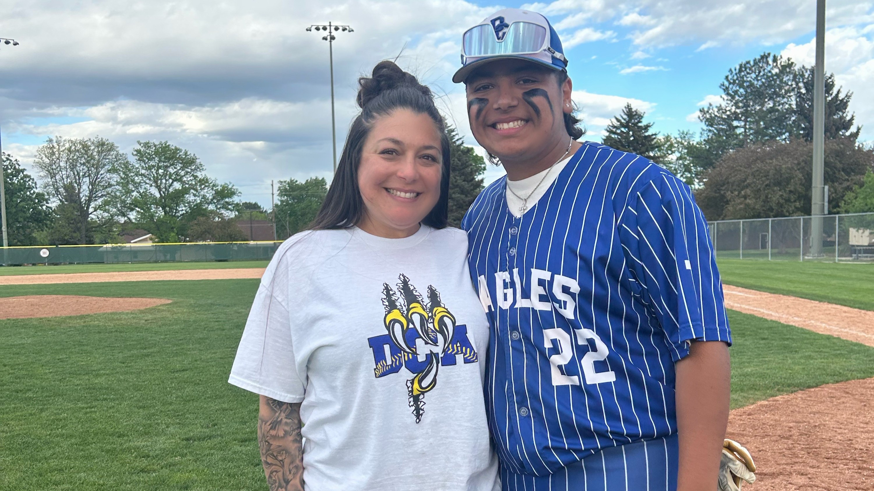 A teenager and his mother standing on a baseball field smiling, one wearing a "DCA" t-shirt and the other in a striped baseball uniform with "Eagles 22" on the front. 