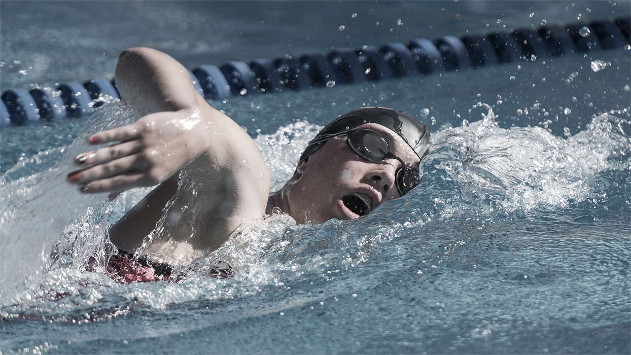 A teenager swims in a lane pool wearing a swim cap and goggles.