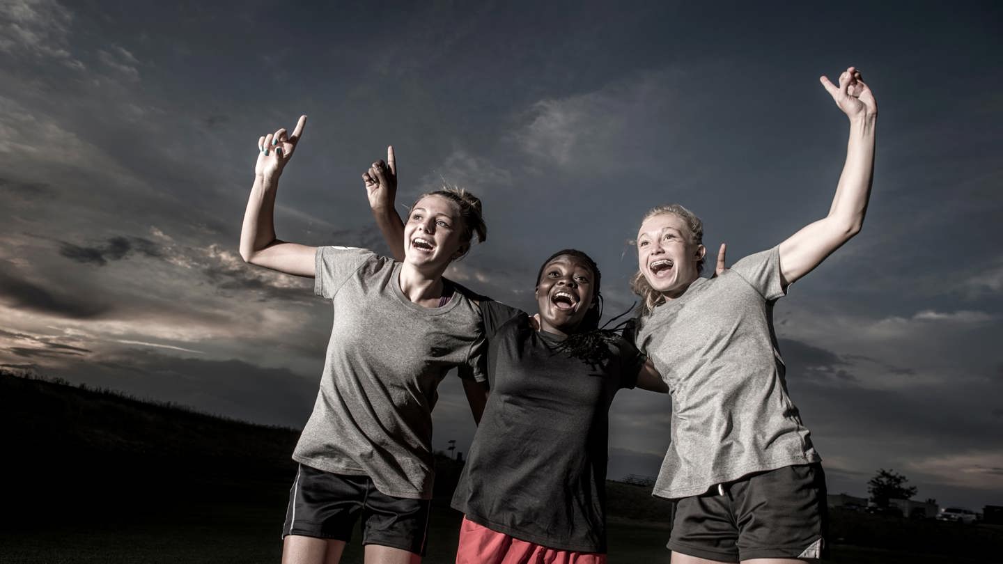 Three teenage female athletes smile and put their arms around each other while holding up "number 1"s with their fingers.