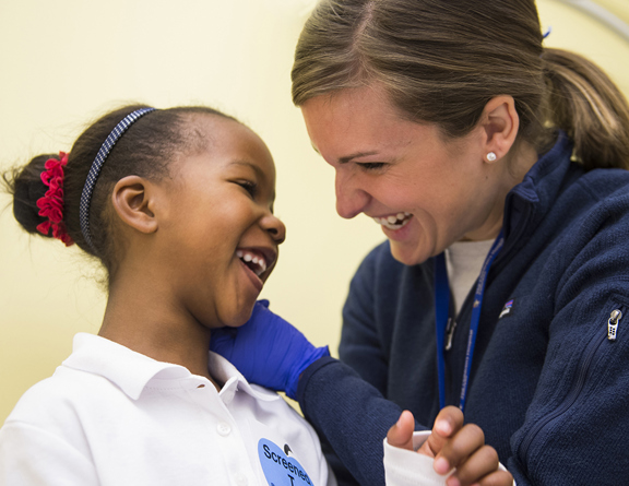 A woman with brown hair and wearing a blue fleece jacket smiles while examining a smiling girl's ear.
