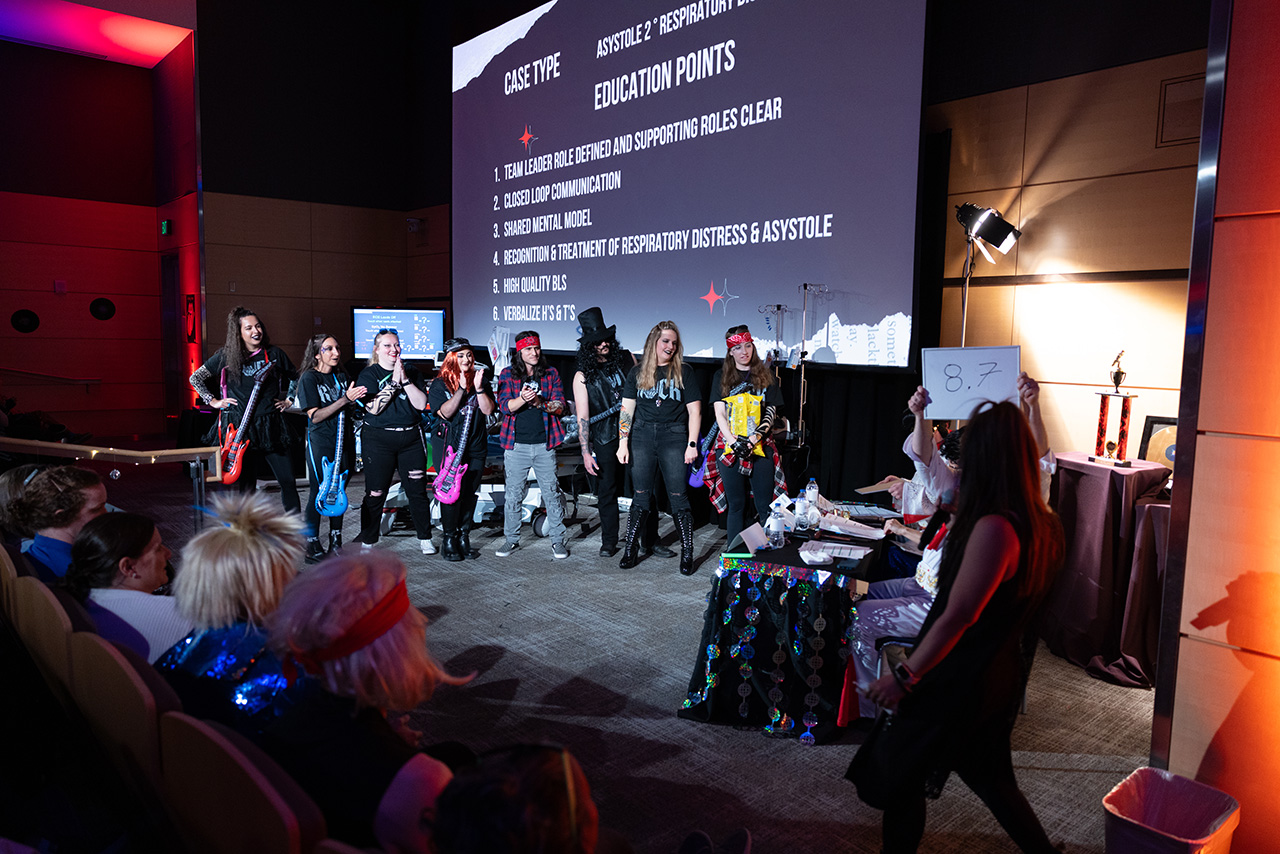 Sim Wars teams stand on stage in front of a large screen listing education points, while a judge holds up a scorecard and an audience watches from theater seating
