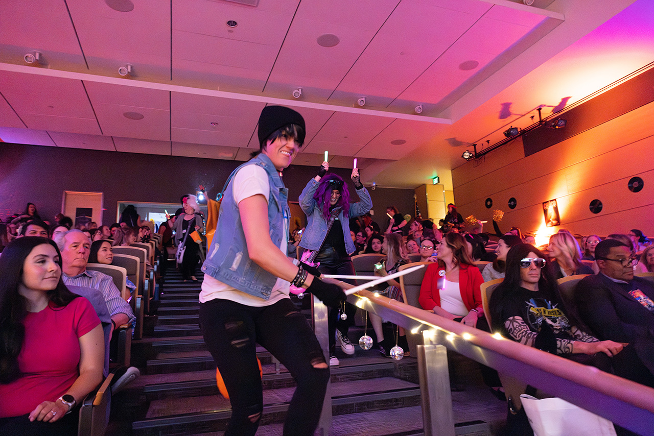 Costumed participants move through an auditorium aisle during Sim Wars, interacting with the audience while teams and spectators watch from seated rows