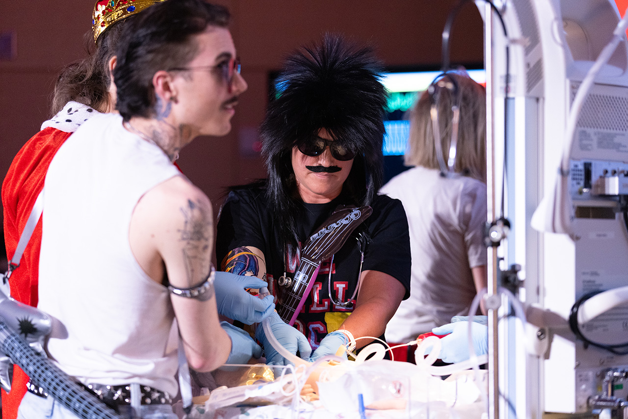 Participants in themed costumes perform a simulated medical resuscitation during Sim Wars, working together around a patient mannequin with clinical equipment and monitors in a training environment