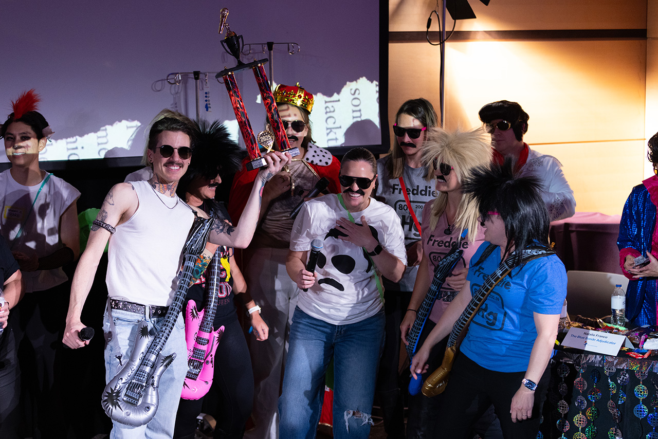 Group of Sim Wars participants in themed rock-style costumes gather on stage, holding inflatable guitars and a large trophy while posing together