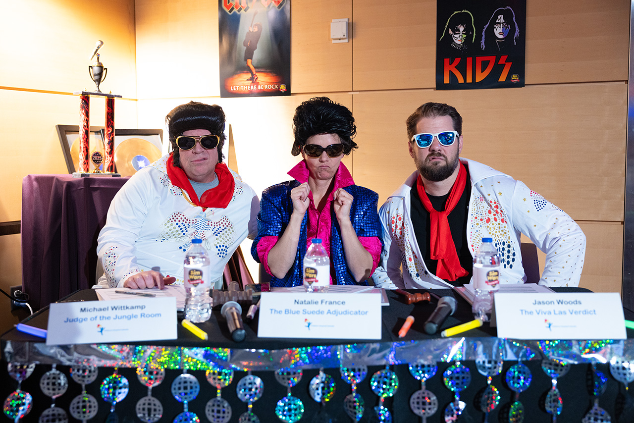 Three Sim Wars judges in coordinated, rock-inspired costumes sit behind a decorated judging table with name placards, microphones, and scoring materials visible, with event trophies and themed posters displayed in the background