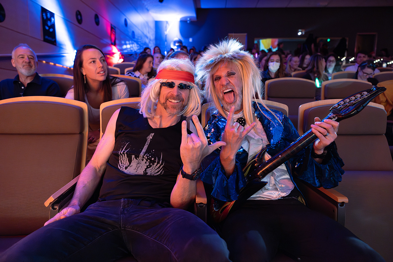 Two Sim Wars attendees in rock-inspired costumes sit in an auditorium audience, posing with hand gestures and a prop guitar 