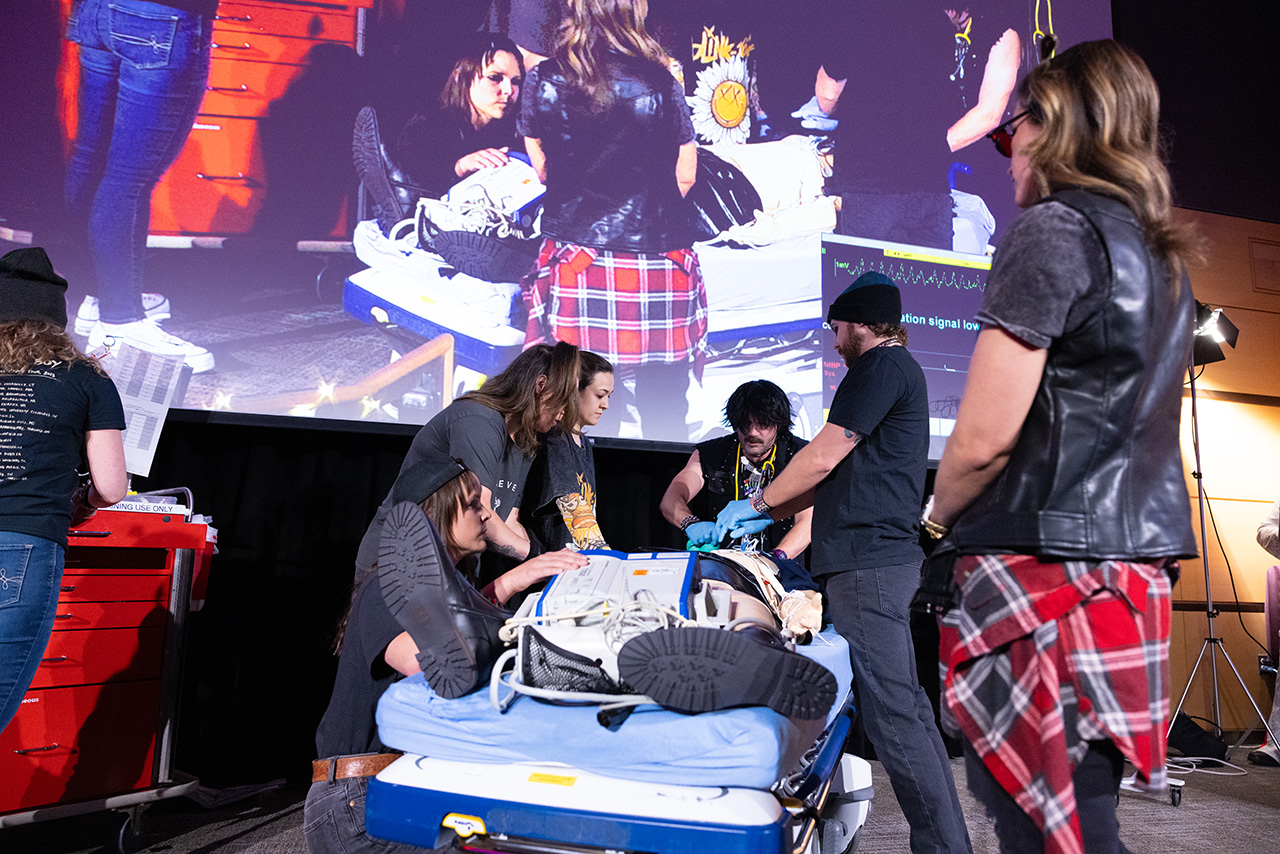 Sim Wars participants in themed costumes perform a simulated patient resuscitation on a hospital bed placed on a stage, while a large screen behind them projects a close-up of the same scene