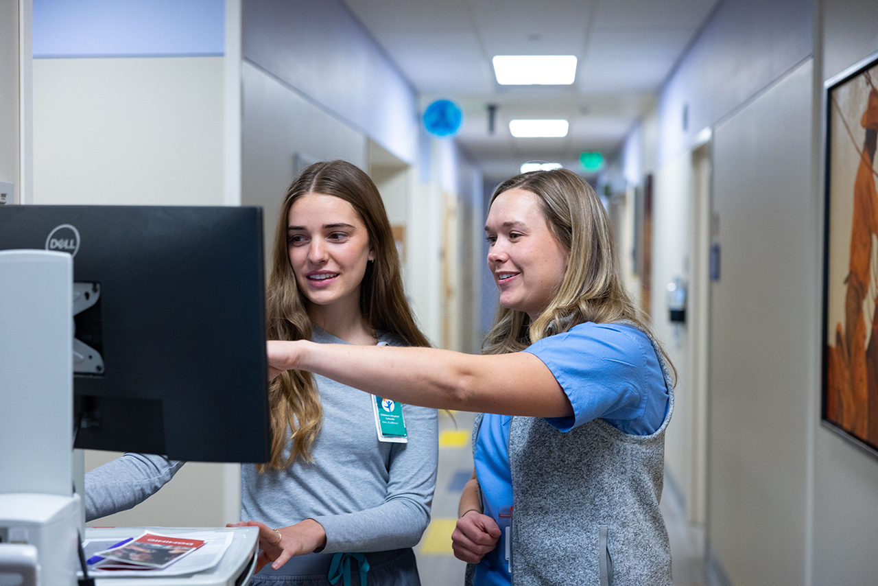 A pediatric nurse in a clinical setting reviewing patient records on a computer monitor.