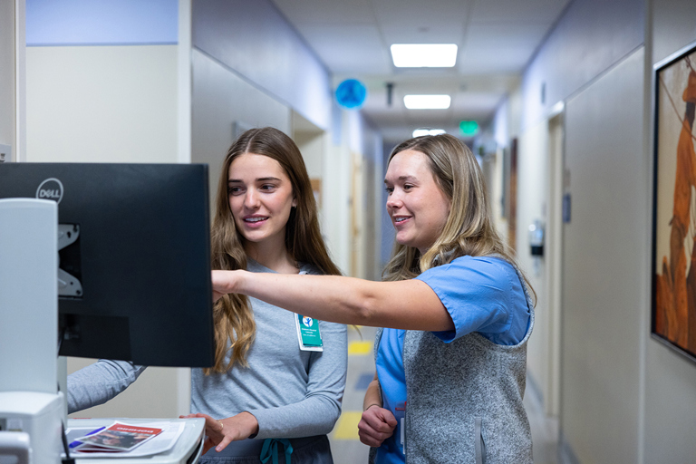 A pediatric nurse in a clinical setting reviewing patient records on a computer monitor.
