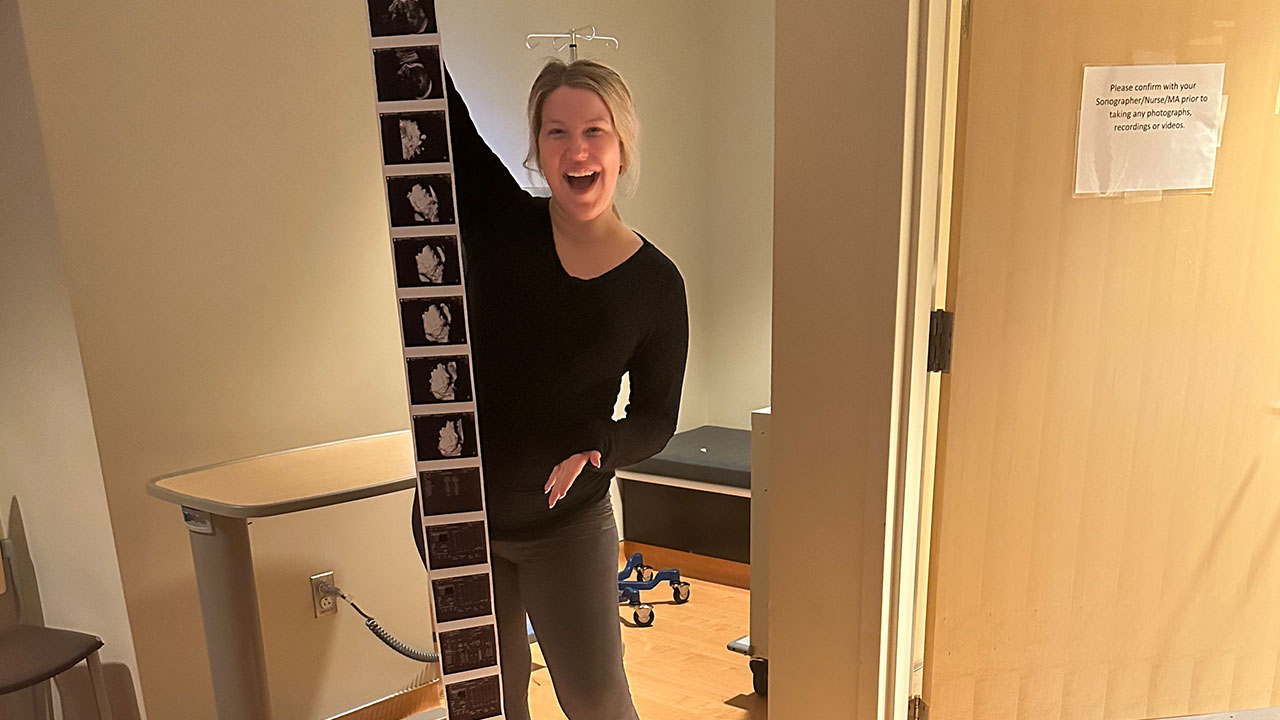 A parent stands smiling in a hospital room with a tall strip of ultrasound images of their baby in front of them.