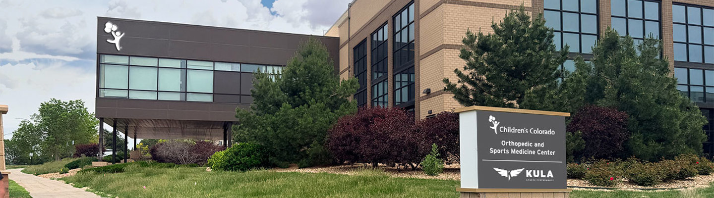 Exterior view of the Children's Colorado Orthopedic and Sports Medicine Center building. The two-story brick and glass building is surrounded by trees and landscaping, with a sign displaying the Children's Colorado and Kula logos.