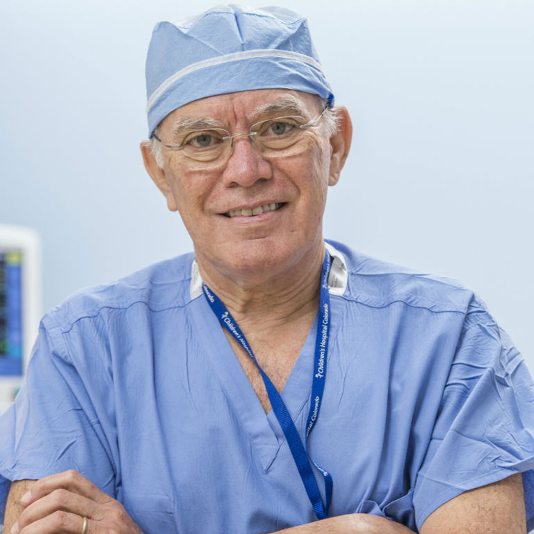 Dr. Alberto Pena smiles at the camera wearing blue scrubs and a surgical cap.