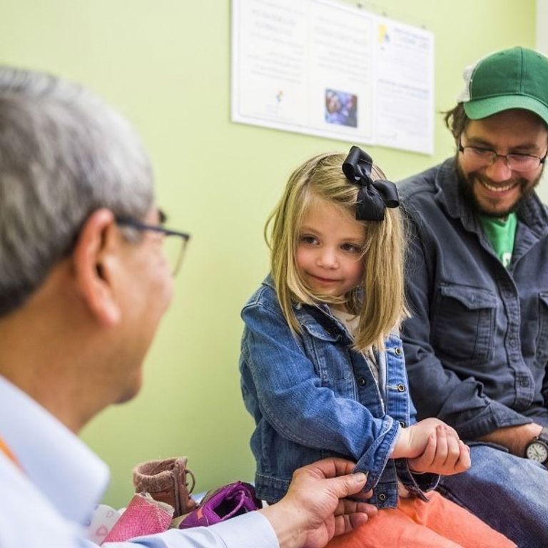 A young girl wearing a denim jacket, peach pants and a black bow in her hair holds her hand and looks at a doctor with glasses and a white lab coat while he tugs on her jacket sleeve. Her dad is behind her wearing a green hat, glasses, green t-shirt, dark gray button down shirt and jeans.