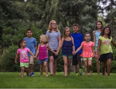 A group of nine kids ranging from toddler to teenager walk in a park holding hands.