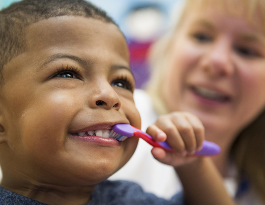 A young boy wearing a blue shirt smiles and brushes his teeth with a purple and pink toothbrush while a doctor with long blonde hair watches in the background.