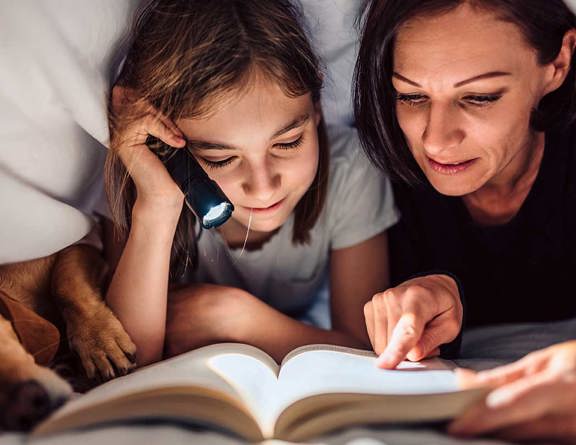 A mom and daughter cuddle with their dog while reading a book under the covers