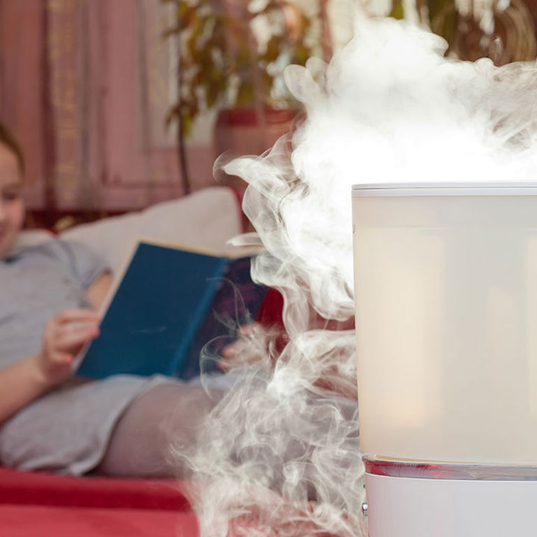 A white humidifier has a smokey vapor coming out of it while a young girl reads a blue book on a red bed with white pillows in the background.