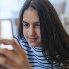 An adolescent girl with brown hair lies on her belly on the couch playing on her smartphone.