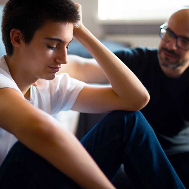 A teen boy and his father sit on the floor in front of a couch. The teen looks upset and his dad is showing support and concern.