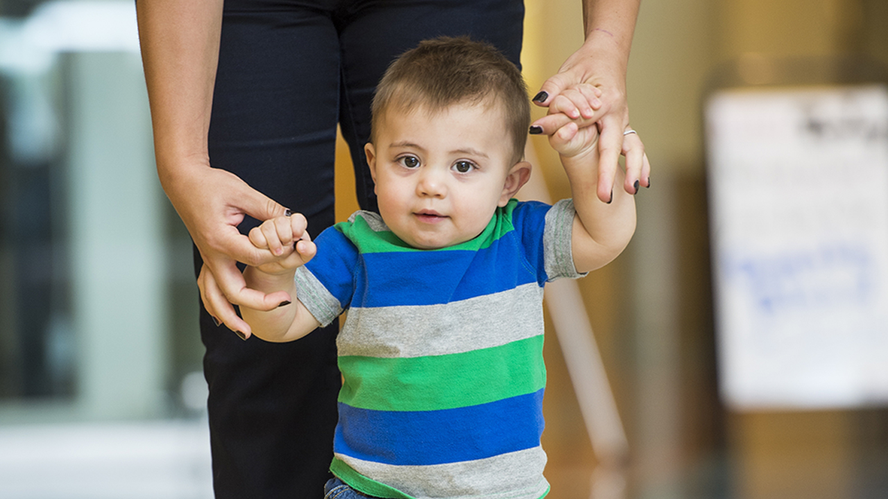 Toddler boy with brown hair walking as he holds on to his mom's hands.