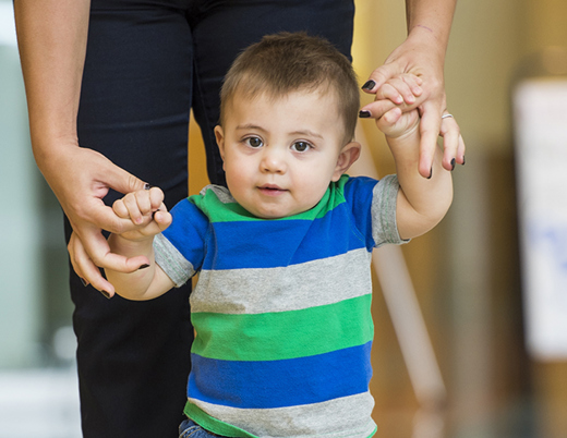 Toddler boy with brown hair walking as he holds on to his mom's hands.