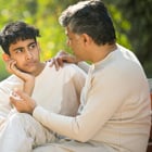 An adolescent boy and his father sit outside together while having a conversation.