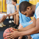 A basketball player sits on a bench in the locker room holding a basketball and looking down while his teammates look happy.