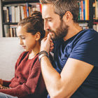 A teenage girl and her dad sit on a couch reading a tablet together.
