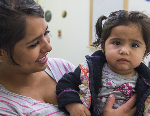 A woman with long brown hair, wearing in a gray sweater and blue latex gloves holds a needle in one hand while holding a boy's arm in the other hand, preparing to give him a shot. The boy has dark hair and is wearing a blue Super Bowl shirt with white and orange lettering.